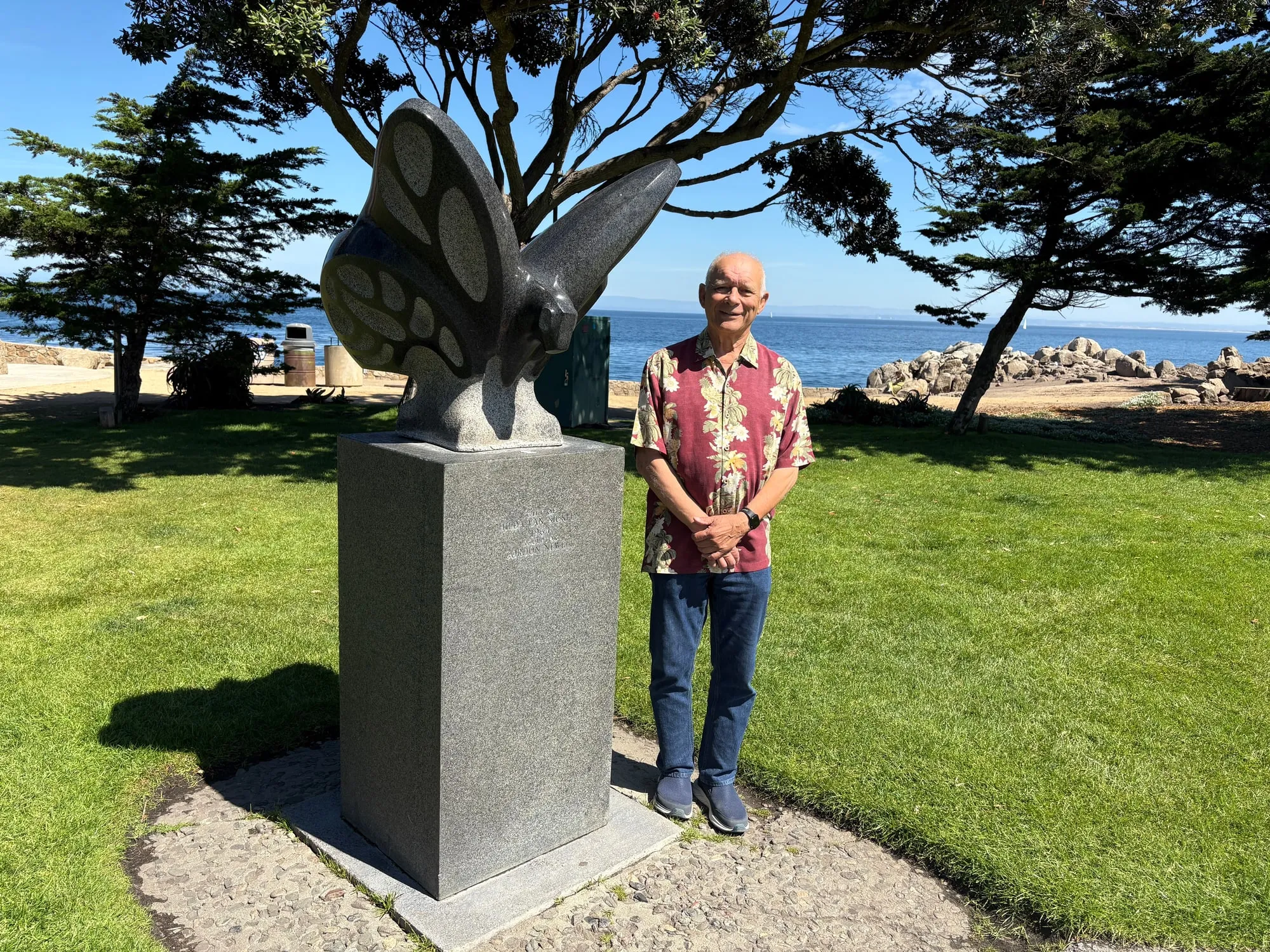 Andrew Kubica standing beside the granite monarch butterfly sculpture in Pacific Grove, with Monterey Bay and a cypress behind him.